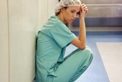 Nurse in scrubs and a hairnet sitting on the floor against a hospital wall, looking stressed and exhausted.