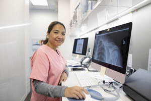 Female healthcare worker in pink scrubs reviewing a chest X-ray on a computer in a medical office.